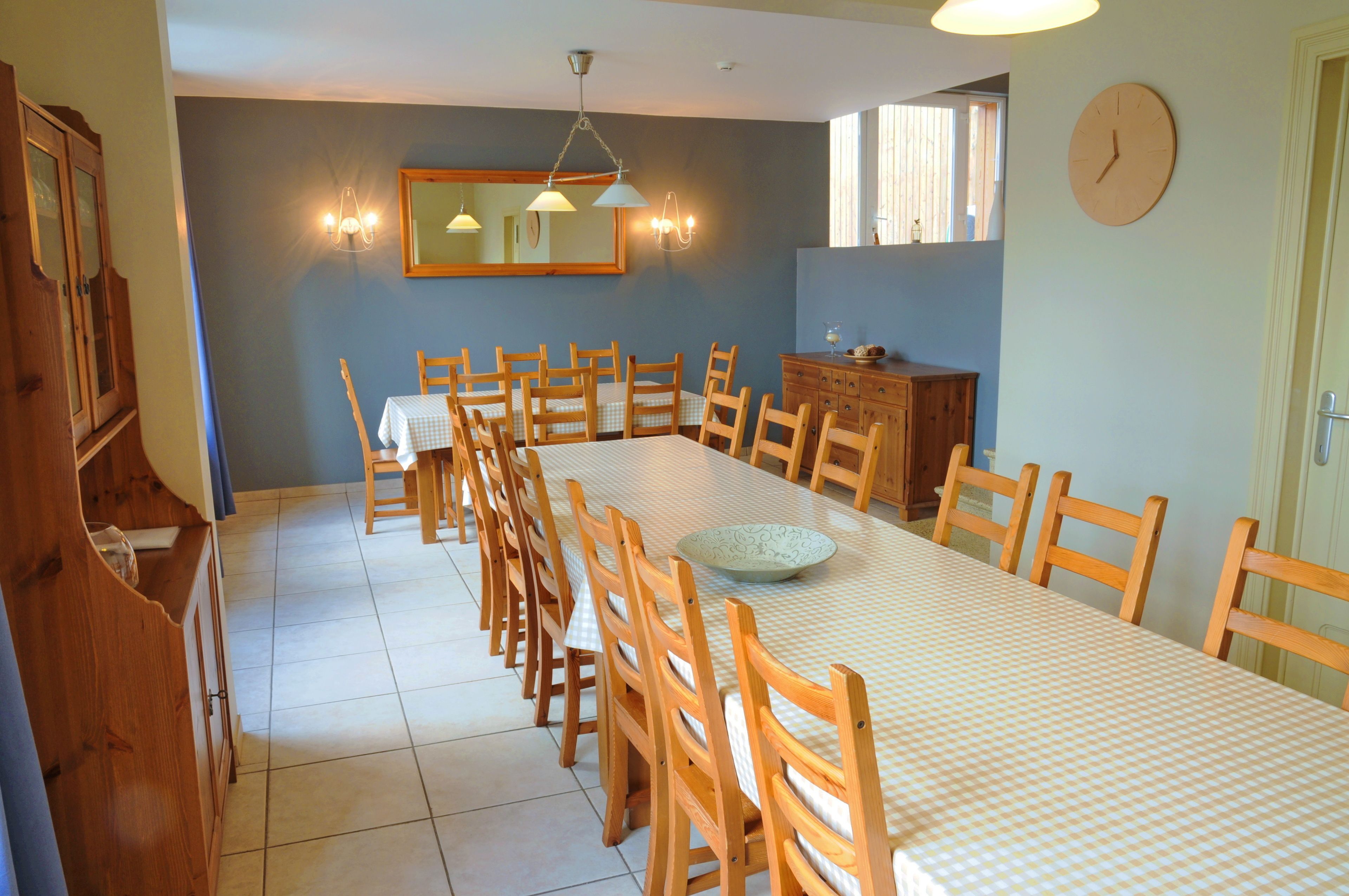 A dining area with a long table, wooden chairs, a checkered tablecloth, a bowl, a mirror, and a clock on the wall.