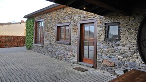 A stone building with a wooden door and windows, a brick pathway, and a metal vent. - Fermette Arbrefontaine (Lierneux)
