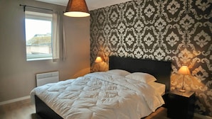 A bedroom with a bed, two bedside lamps, a window with curtains, and a patterned wallpaper.