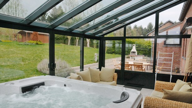 Zona de hidromasaje con techo de cristal, mobiliario de mimbre y vistas a un jardín y una casa.