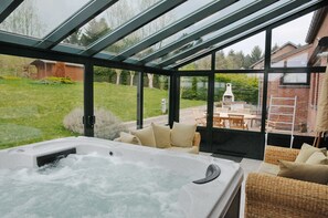 Een bubbelbadgedeelte met glazen dak, rieten meubels en uitzicht op tuin en huis.