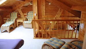 Una habitación acogedora en el ático con vigas de madera, dos sillones de mimbre con cojines a cuadros azules y amarillos, una mesita y una cama morada.