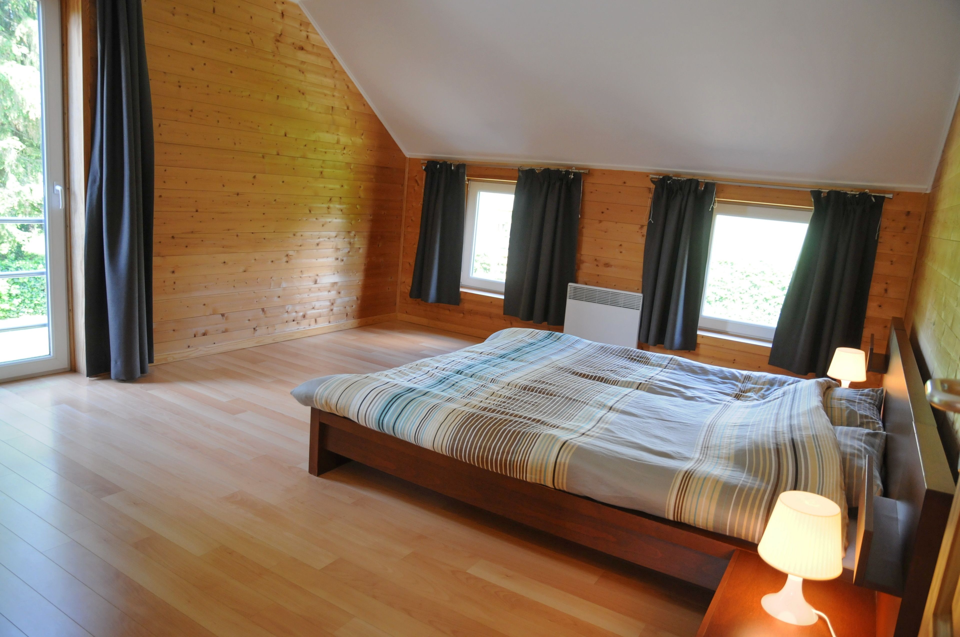 A bedroom with a wooden floor, a bed with a striped bedspread, two windows with curtains, and a nightstand with a lamp.