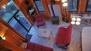A cozy cabin interior with wooden walls, a fireplace, red seating, and large windows offering a view of the outdoors. - Jardins de l'Ourthe 16 (Houffalize)