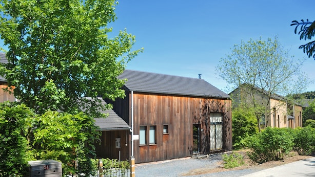 A wooden building with a metal door, surrounded by trees and a gravel driveway. - Jardins de l'Ourthe 16 (Houffalize)