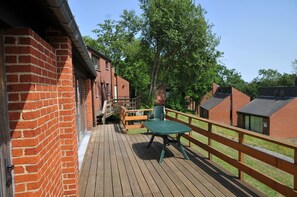 A wooden deck with a green table and chairs, surrounded by brick buildings and trees.