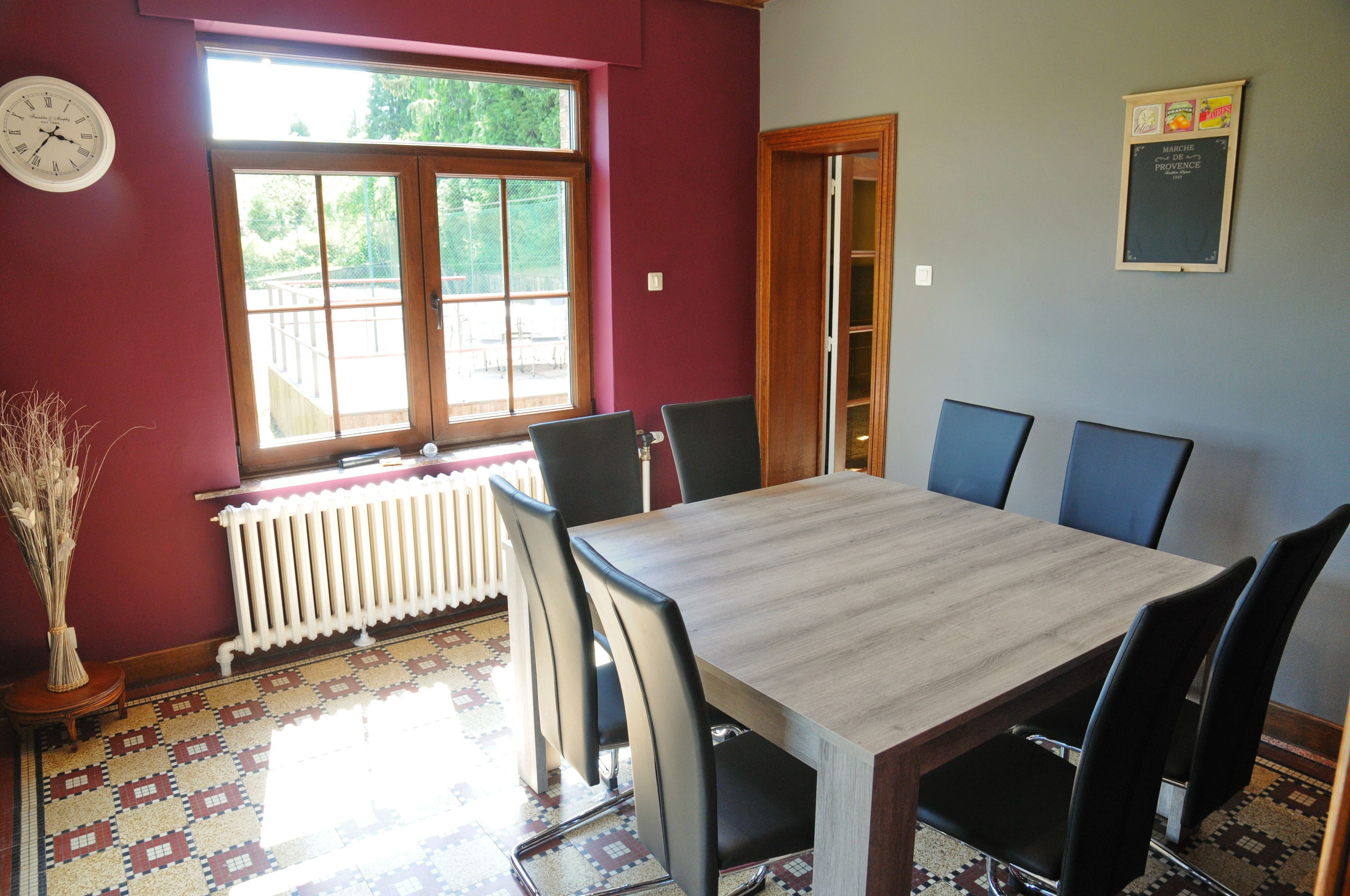 A dining room with a table and chairs, a window with a view, a radiator, and a clock on the wall.