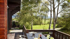 A wooden deck with a table set for dining, surrounded by chairs, and overlooking a grassy landscape with trees.