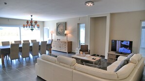 Ein modernes Wohnzimmer mit einem großen weißen Sofa, einem Essbereich mit Stühlen, einer Kristallleuchter, einer Uhr und einem Fernseher.