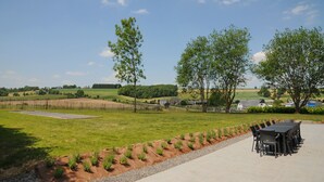 Ein Essbereich im Freien mit einem langen Tisch und Stühlen, umgeben von einem angelegten Garten mit Bäumen und Blick auf die Landschaft.