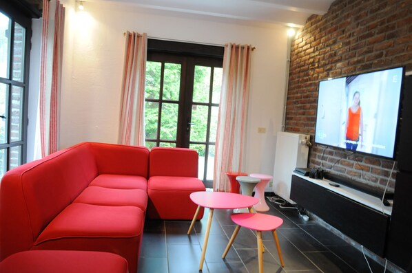 A modern living room with a red sectional sofa, a flat-screen TV mounted on a brick wall, and a large window with curtains.