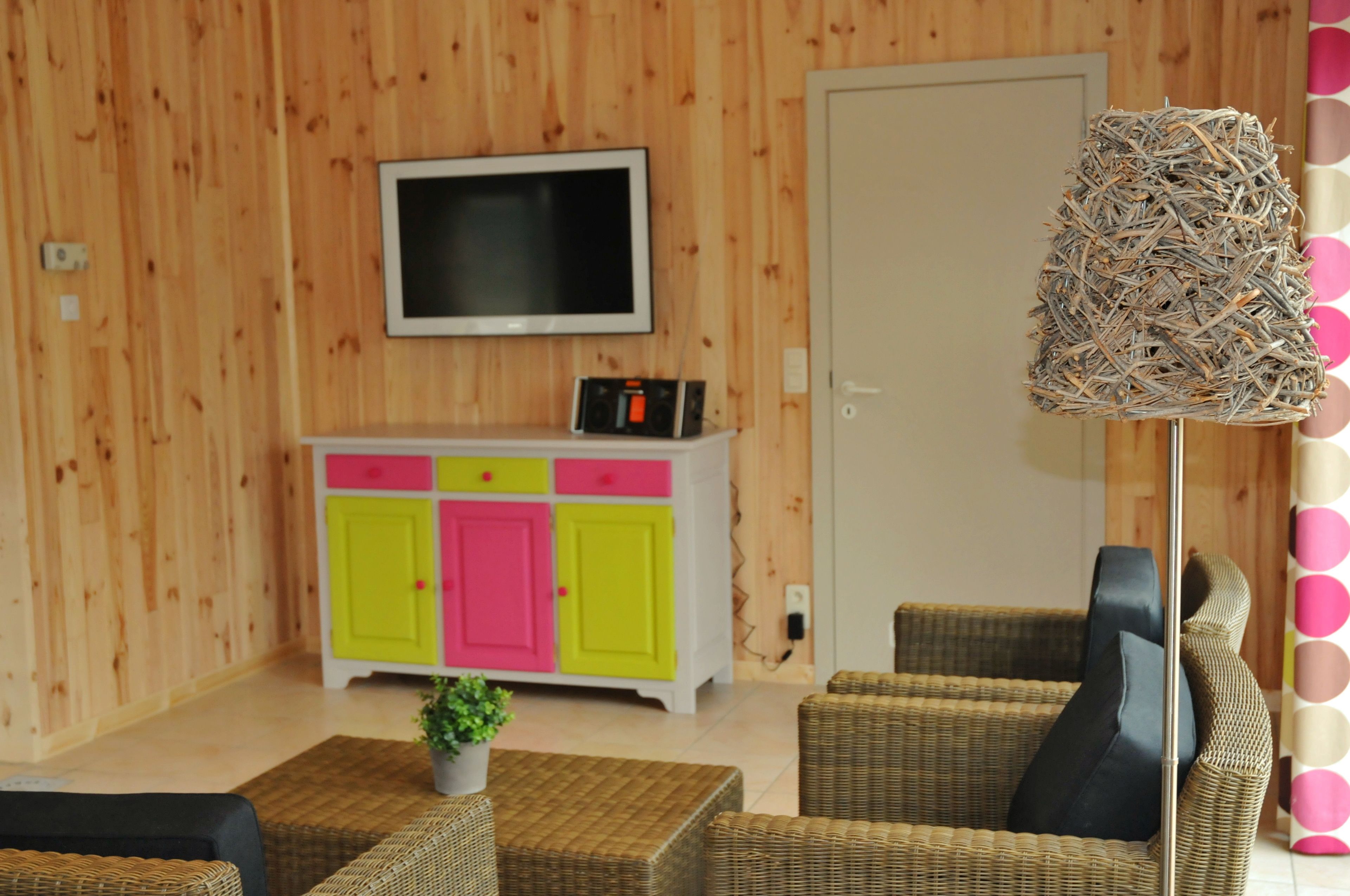 A room with a television mounted on the wall, a colorful cabinet, wicker furniture, and a decorative lamp.