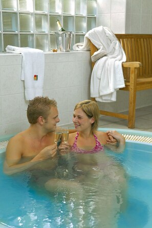 A man and a woman in a hot tub with champagne glasses, surrounded by towels and a wooden chair. - Résidence Thier Antoine (Somme-Leuze)