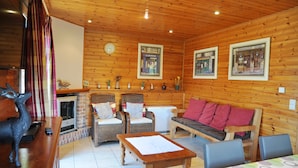 A cozy cabin interior with wooden walls, a fireplace, wicker furniture, and framed pictures on the walls. - Rochettes - Pluton 3 (La Roche-en-Ardenne)