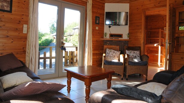A wooden cabin interior with a sofa, two chairs, a table, a TV mounted on the wall, and a sliding glass door leading to an outdoor deck. - Rochettes - Vénus 2 (La Roche-en-Ardenne)