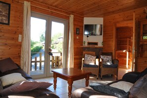 A wooden cabin interior with a sofa, two chairs, a table, a TV mounted on the wall, and a sliding glass door leading to an outdoor deck. - Rochettes - Vénus 2 (La Roche-en-Ardenne)