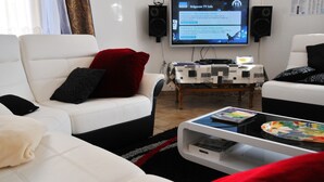 Uma sala de estar moderna com sofás de couro branco, uma televisão de tela plana, caixas de som e uma mesa de centro com livros e um prato.