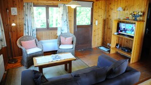 Une pièce chaleureuse aux murs en bois, avec un canapé, deux fauteuils en osier dotés de coussins, une petite table, un téléviseur posé sur une étagère et une fenêtre avec des rideaux.
