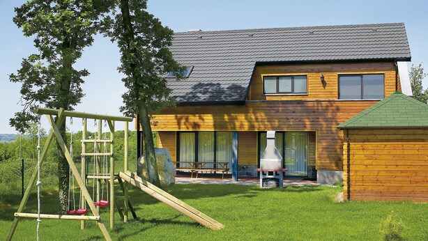 A modern two-story house with a wooden exterior, a green roof, and a swing set in the yard.