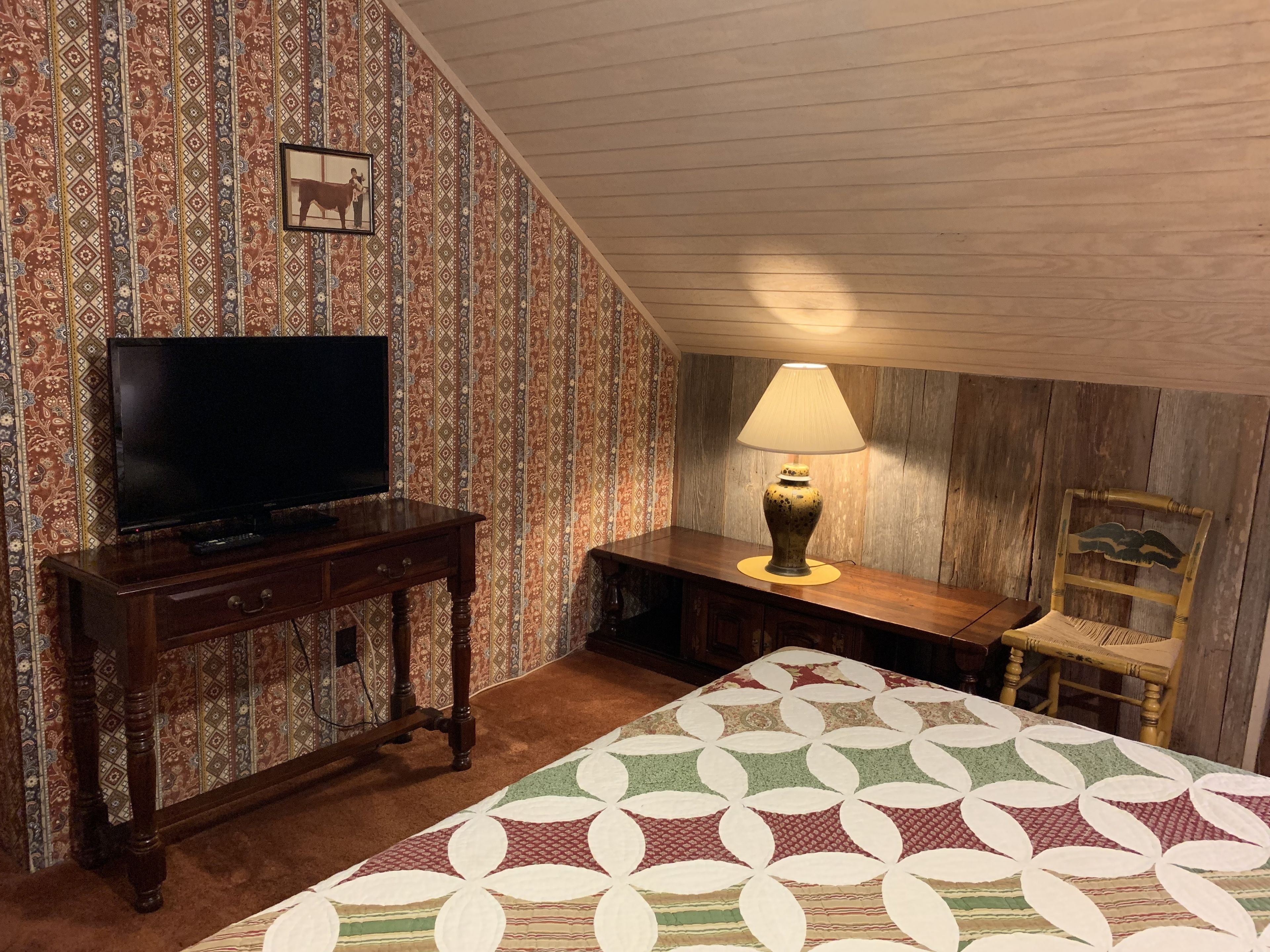 A room with a television on a wooden stand, a lamp on a small table, a bed with a patterned quilt, and a chair with a wooden headboard.