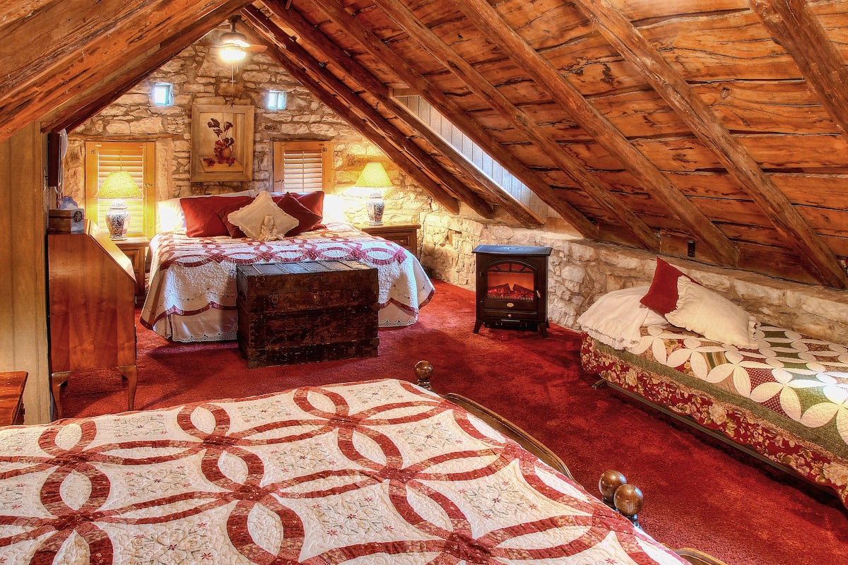 Un dormitorio acogedor en el ático, con techo de madera, pared de piedra, cama con colcha roja y blanca, un cofre de madera y un pequeño estufa de leña.