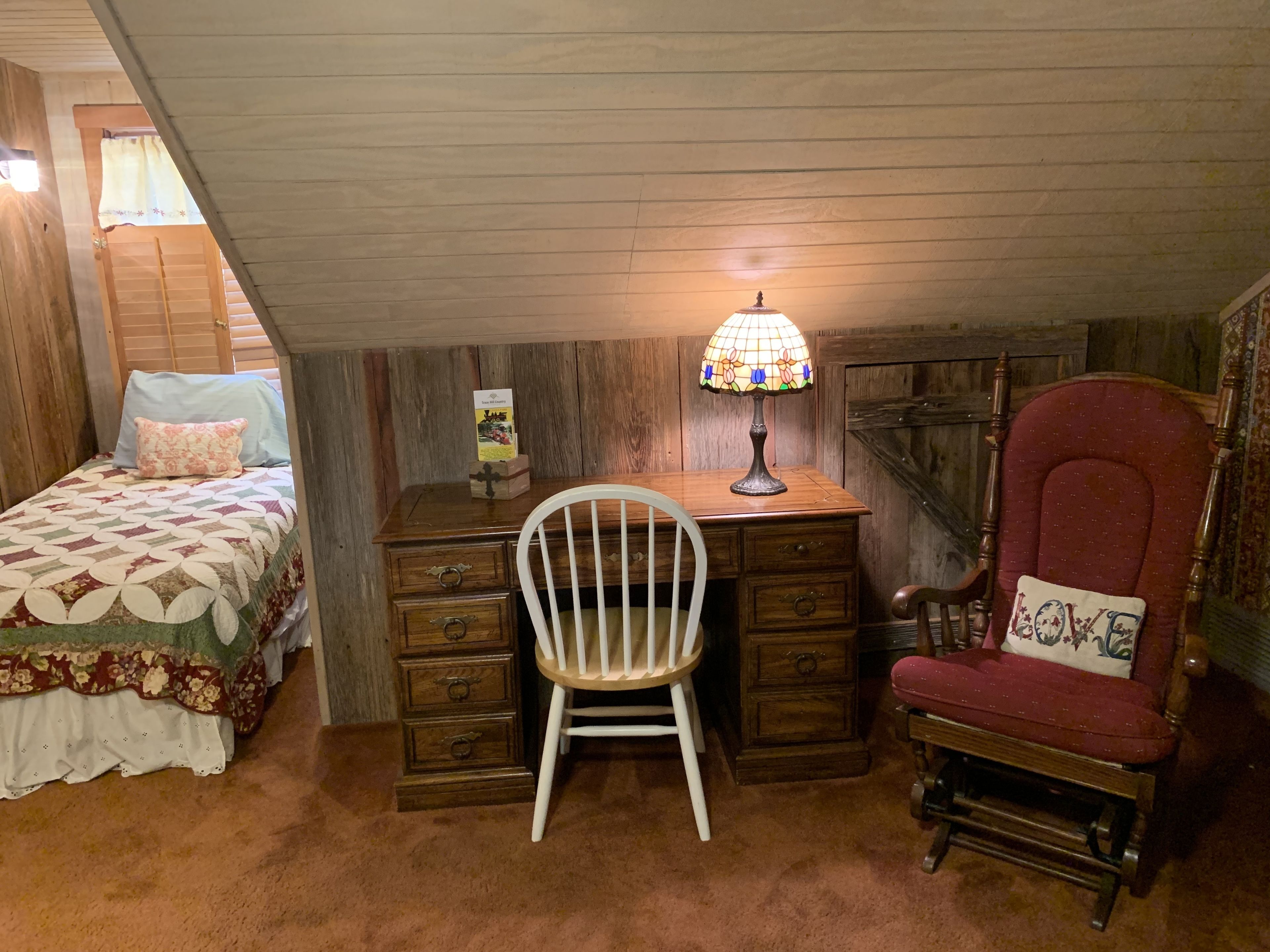 A cozy attic bedroom with a wooden desk, a white chair, a red armchair, a bed with a floral bedspread, and a stained glass lamp.