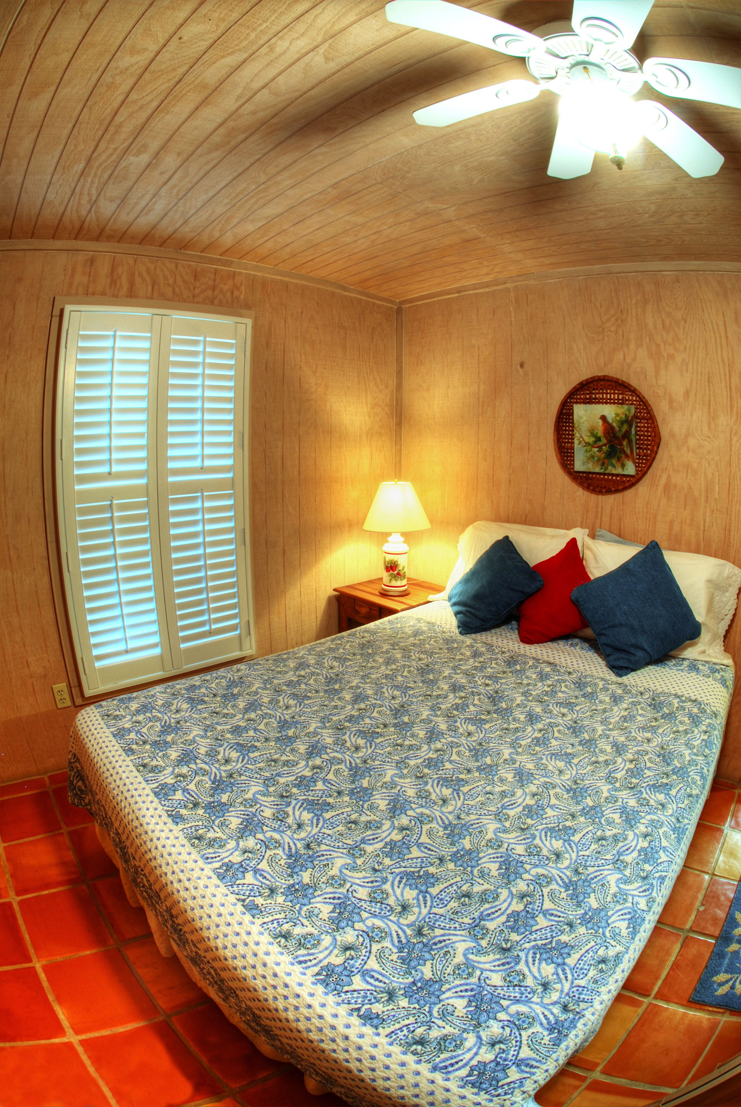 A bedroom with a bed, a ceiling fan, a window with shutters, a lamp, and a wall decoration.