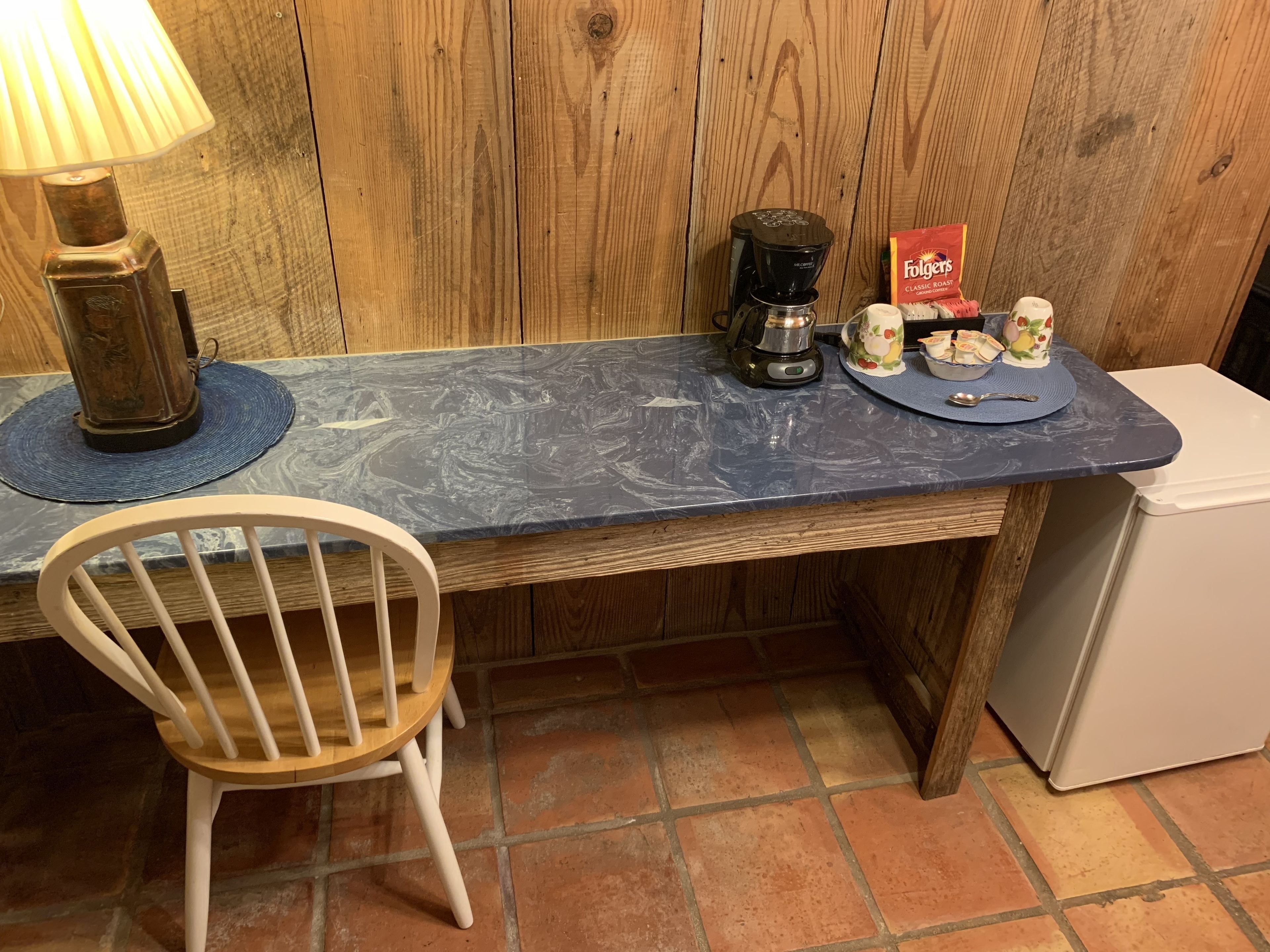 A kitchen with a wooden table, a white chair, a coffee maker, a box of Folgers coffee, and a refrigerator.