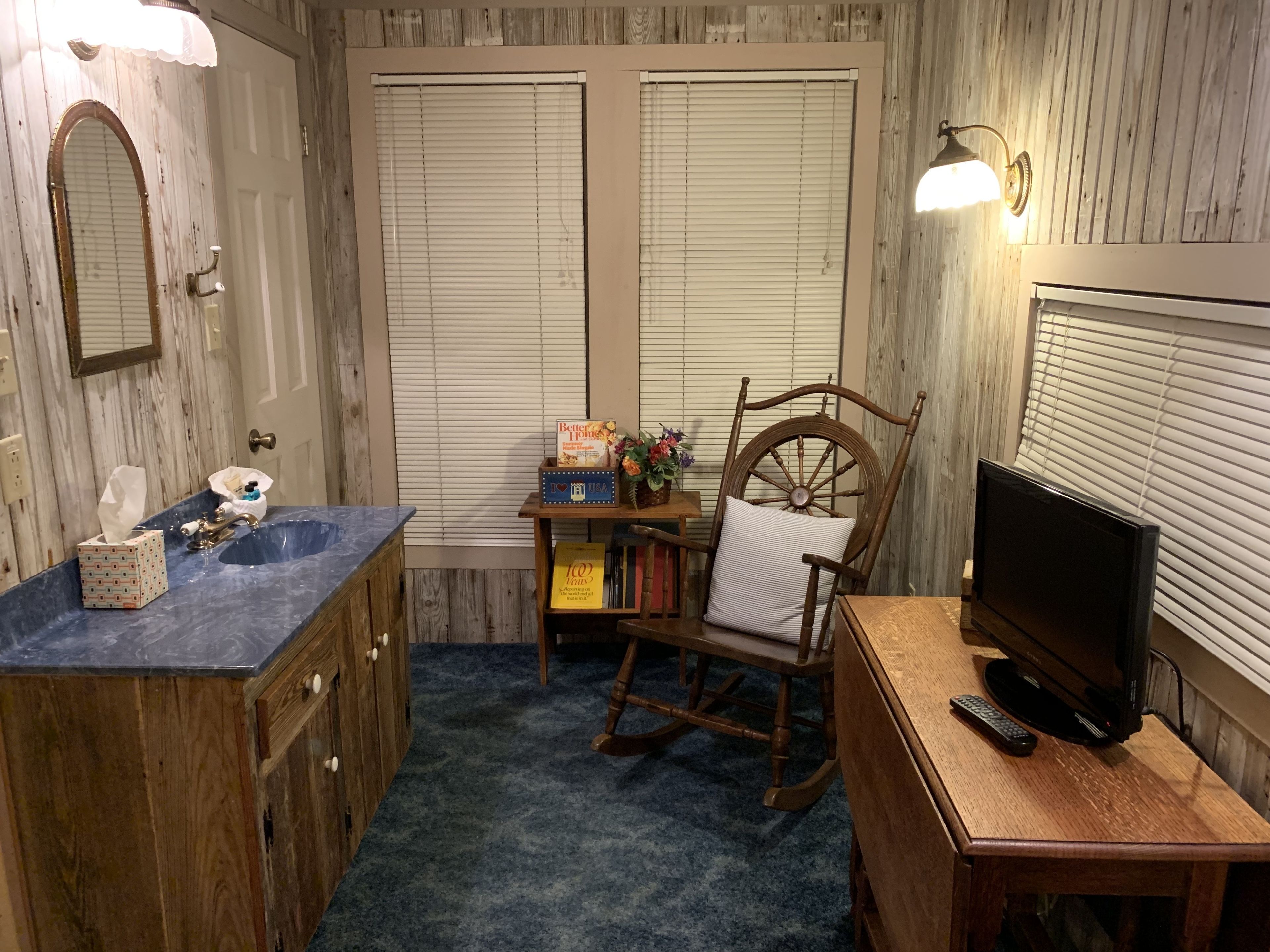Un baño con tocador de madera, un lavabo, un espejo, una mecedora, un televisor y una pequeña mesa con libros y una caja.