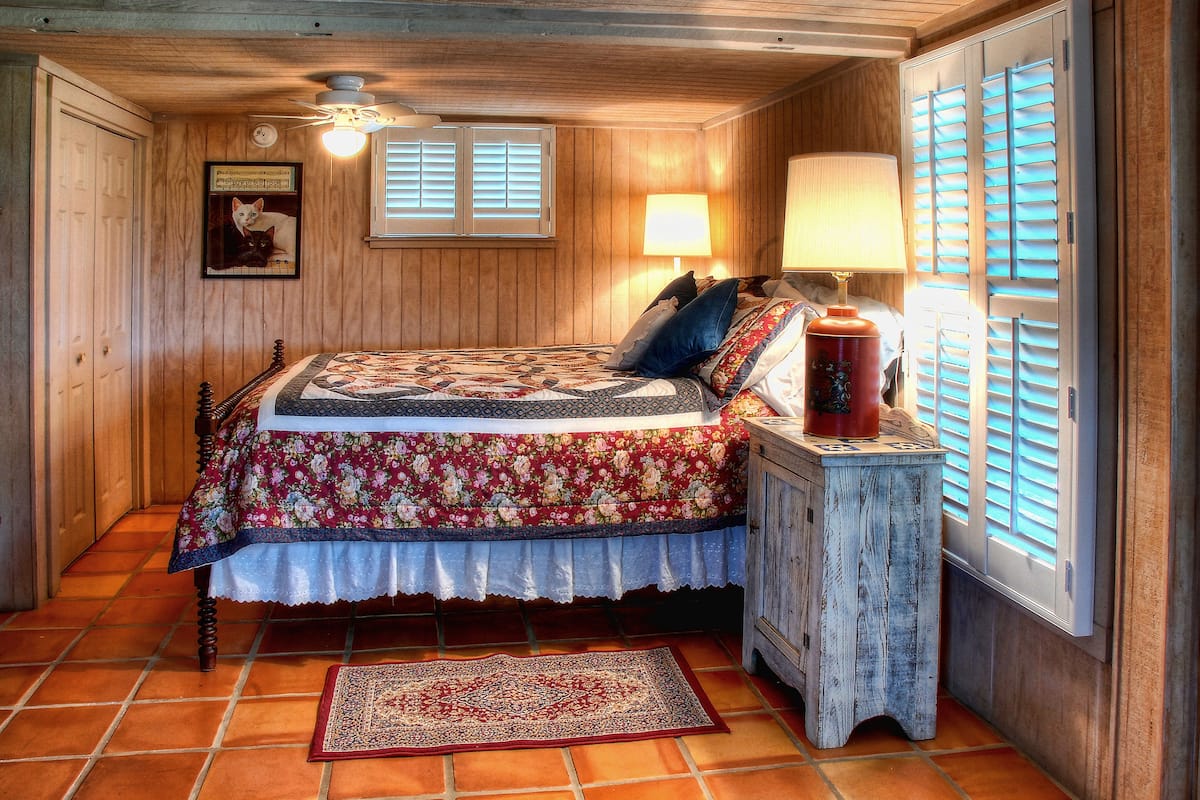 Un dormitorio con techo de madera, una cama con colcha estampada, una mesita de noche con lámpara y una ventana con contraventanas.