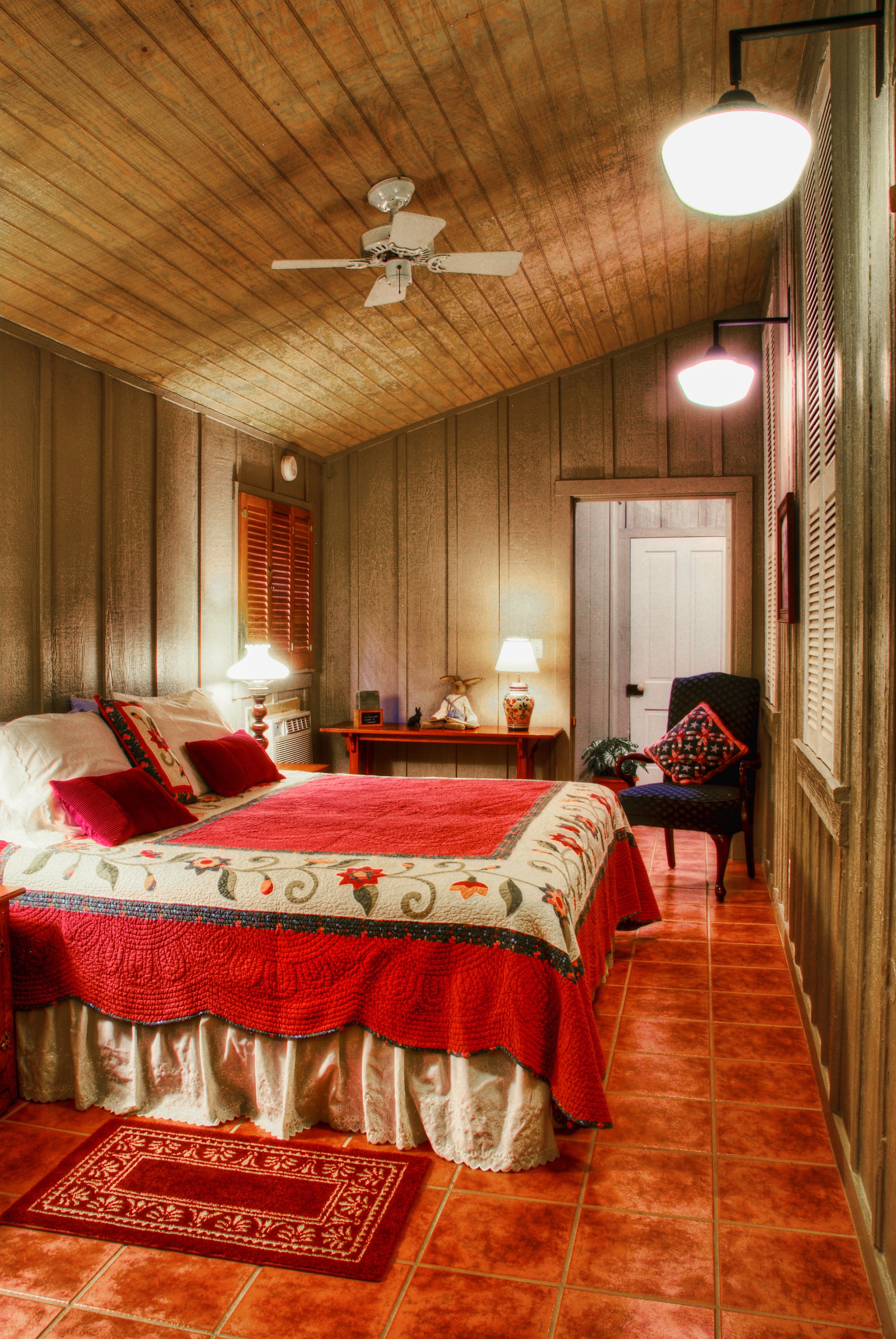 Una habitación con techo de madera, cama con ropa de cama roja y blanca, una silla, una mesa con lámpara y una puerta.