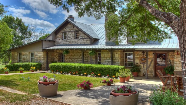 A stone house with a metal roof, a front porch with chairs, and a well-maintained garden with potted plants and flowers.