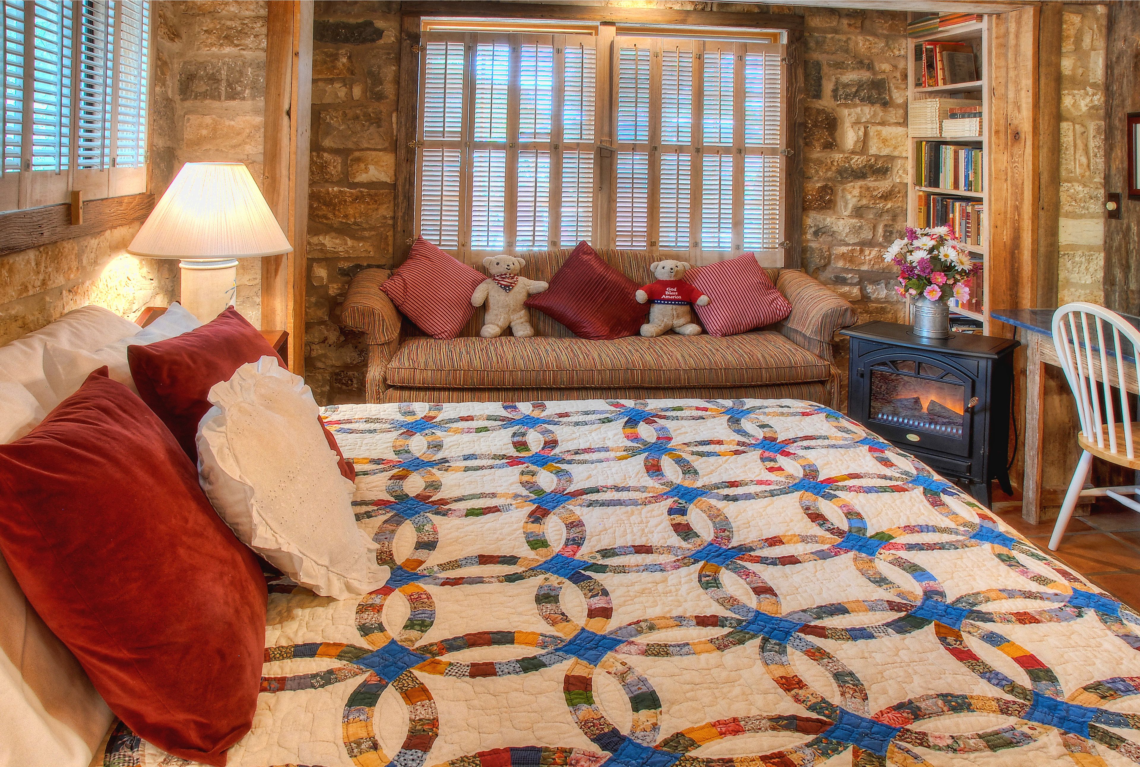 A cozy room with a large bed, a wooden bench with pillows, a quilt, a bookshelf, and a fireplace.