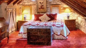 A rustic attic bedroom with exposed wooden beams, a stone wall, a bed with a quilt, a wooden chest, and a nightstand with a lamp.