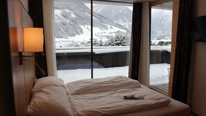 Quarto com uma cama, mesa de cabeceira, abajur e vista para montanhas nevadas através de grandes janelas.
