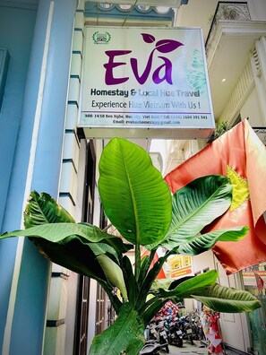 A signboard for 'Eva Homestay & Local Hue Travel' with contact details and a large green plant in the foreground.