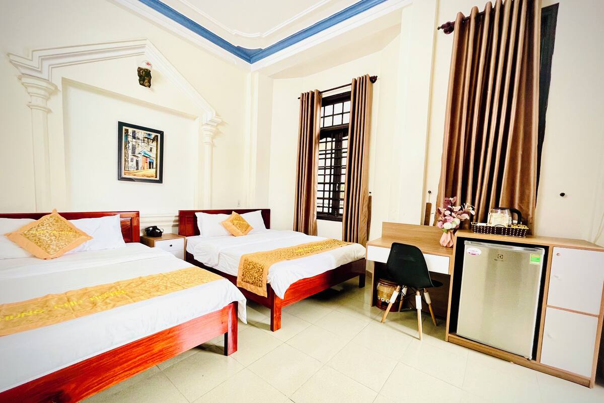 A hotel room with two beds, a desk, a refrigerator, and a window with curtains.