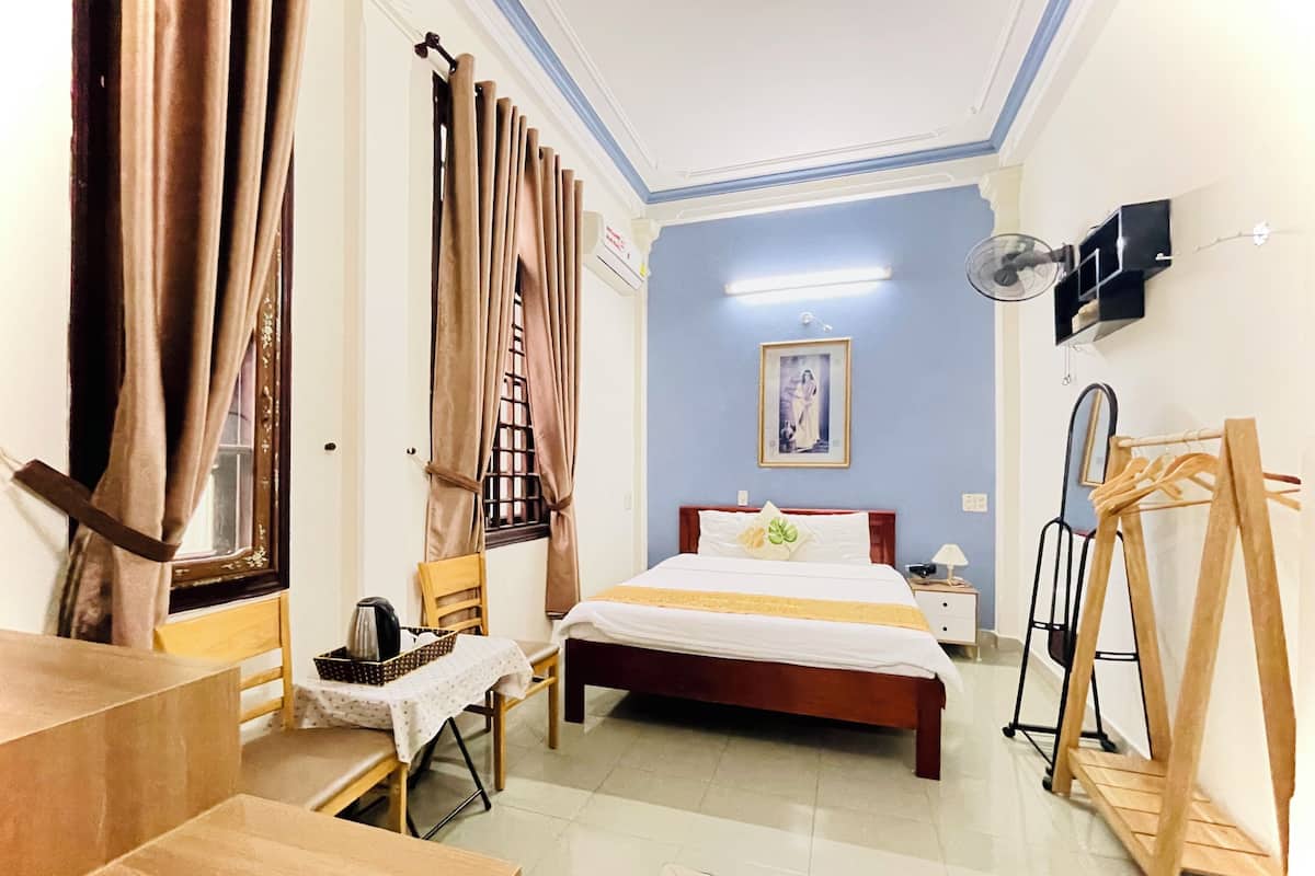A hotel room with a bed, a table, a chair, a fan, and a wooden easel.