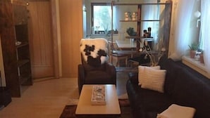 Un salon avec un canapé de couleur foncée, une table basse et un fauteuil recouvert d’un plaid à motifs. On y trouve également une bibliothèque et une fenêtre avec des rideaux.
