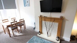 Een woonkamer met een open haard, een televisie erboven bevestigd, een eettafel met stoelen, en een blauw vloerkleed.