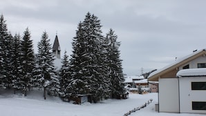 Paisaje nevado con una iglesia, altos árboles de hoja perenne y un edificio moderno con techo inclinado.
