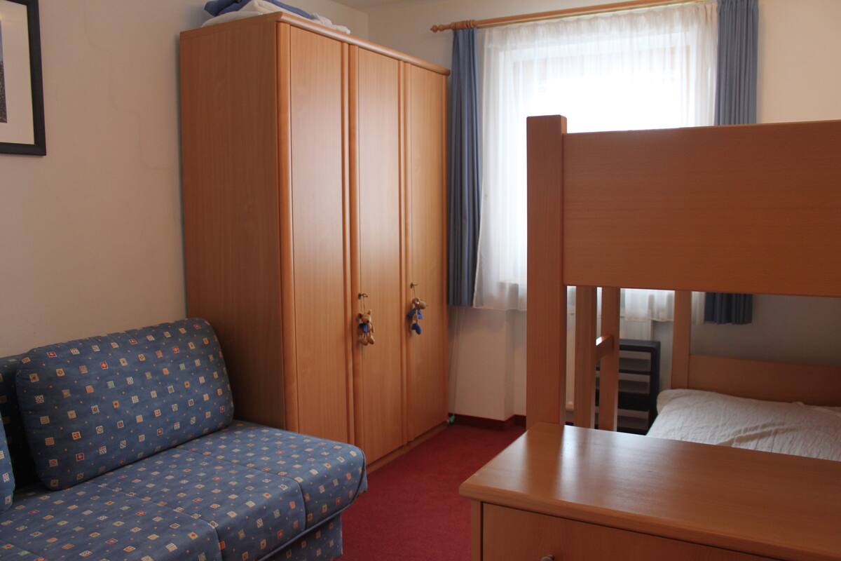 A room with a bunk bed, a wardrobe, a sofa, and a window with curtains.