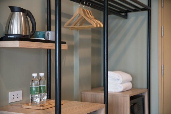 A hotel room with a wooden shelf holding a kettle, cups, and water bottles. There is a wooden hanger and a folded towel on a wooden table.