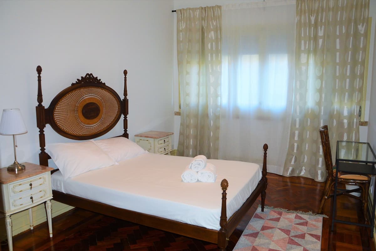 A neatly made bed with white linens and rolled towels, a wooden headboard, a bedside table with a lamp, and a chair in a room with wooden flooring and a patterned rug.