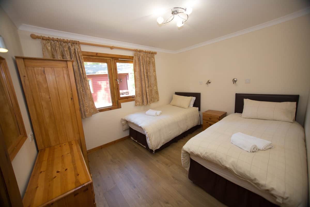 A bedroom with two beds, wooden flooring, a window with curtains, and a ceiling light fixture.