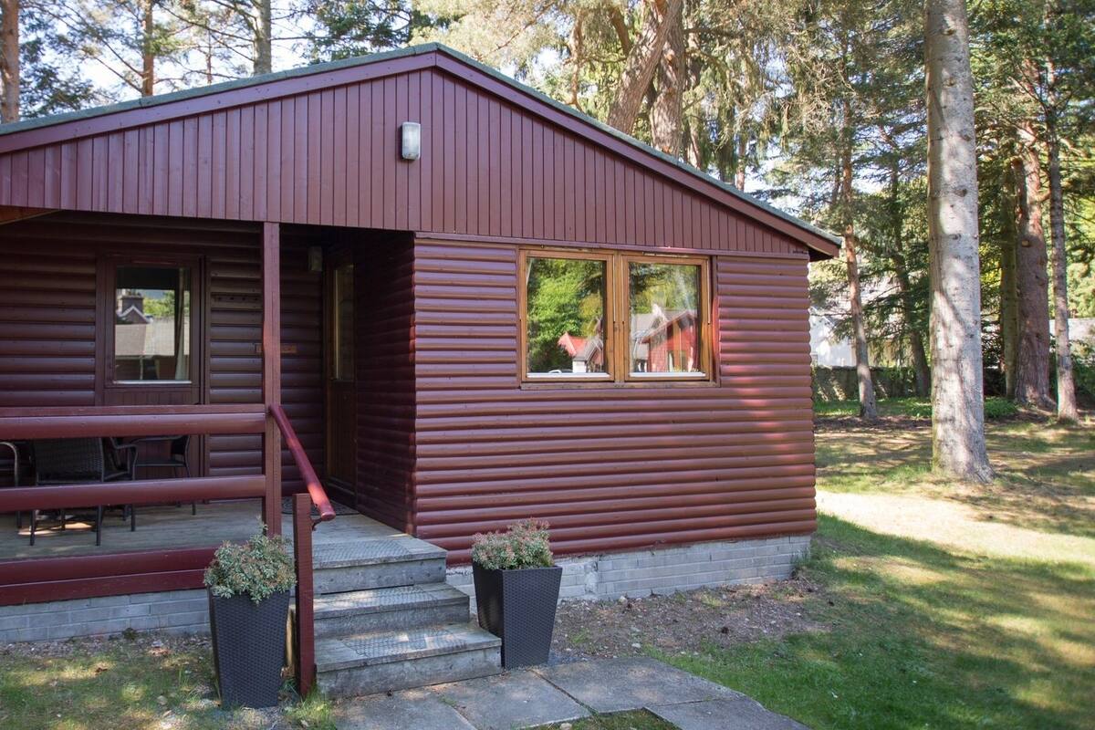 A small, single-story cabin with a porch and a small deck, surrounded by trees.