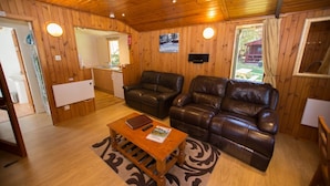 A cozy living room with wooden walls and ceiling, a brown leather sofa, a wooden coffee table with a book and remote, and a kitchen area in the background. - Invercauld Lodges (Ballater)