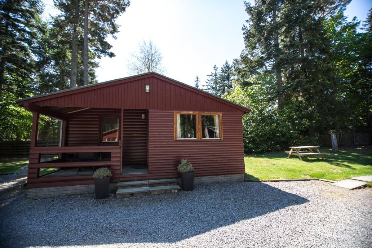 A small, single-story wooden cabin with a porch, a picnic table, and a gravel driveway.