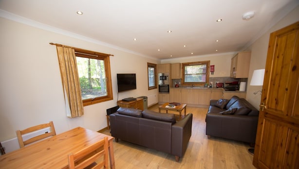 A spacious living area with a wooden dining table, two sofas, a kitchen with wooden cabinets, and large windows. - Invercauld Lodges (Ballater)