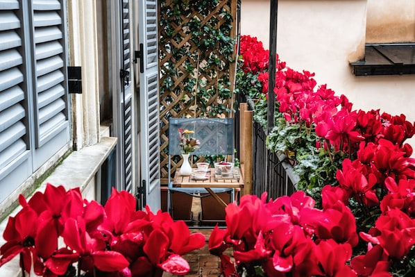 A narrow outdoor corridor with red flowers, a wooden table with a vase, and a lattice wall. - Navona Grand Suite (Rome)